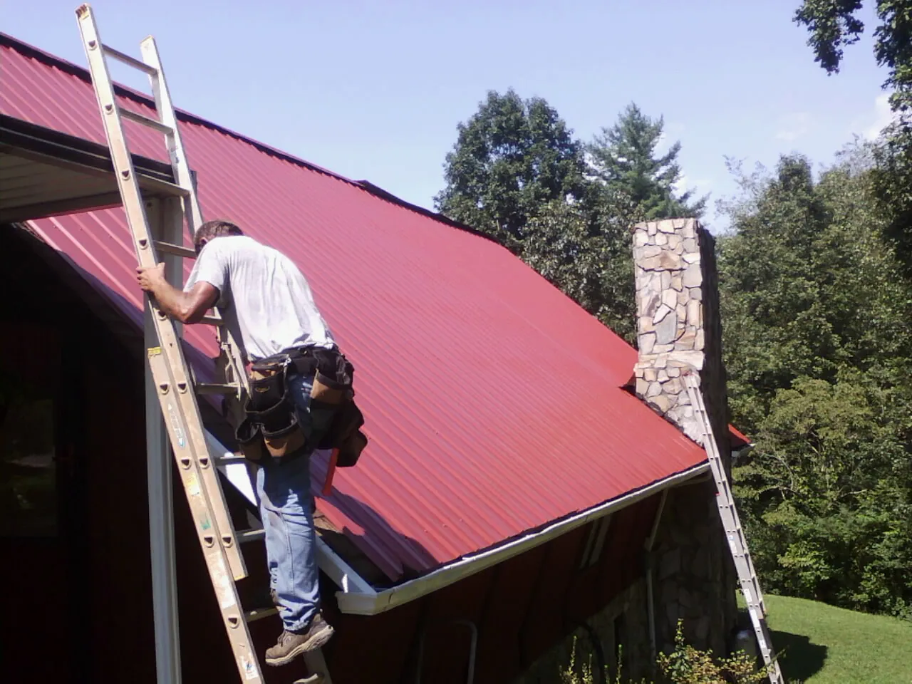 Roofer on a ladder beside a red metal roof and stone chimney — Soffit & Fascia Repair by Roofing Brothers in Sycamore
